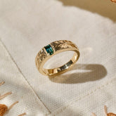 A solid gold ring with an intricate leaf-patterned hand engraved band and a rectangular green blue natural sapphire, resting on a textured cream-colored fabric.