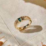 A solid gold ring with an intricate leaf-patterned hand engraved band and a rectangular green blue natural sapphire, resting on a textured cream-colored fabric.