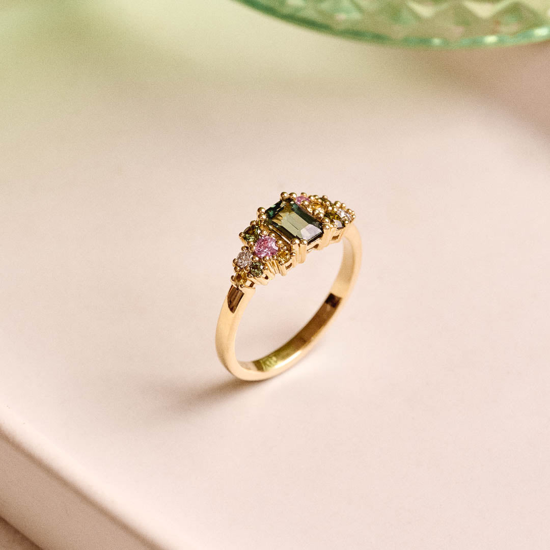 Handmade cluster engagement ring in 9ct yellow gold with a central emerald-cut green sapphire, surrounded by green, yellow, and pink sapphires and two natural diamonds in a floral-inspired design.