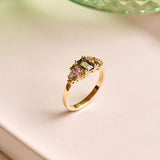 Handmade cluster engagement ring in 9ct yellow gold with a central emerald-cut green sapphire, surrounded by green, yellow, and pink sapphires and two natural diamonds in a floral-inspired design.