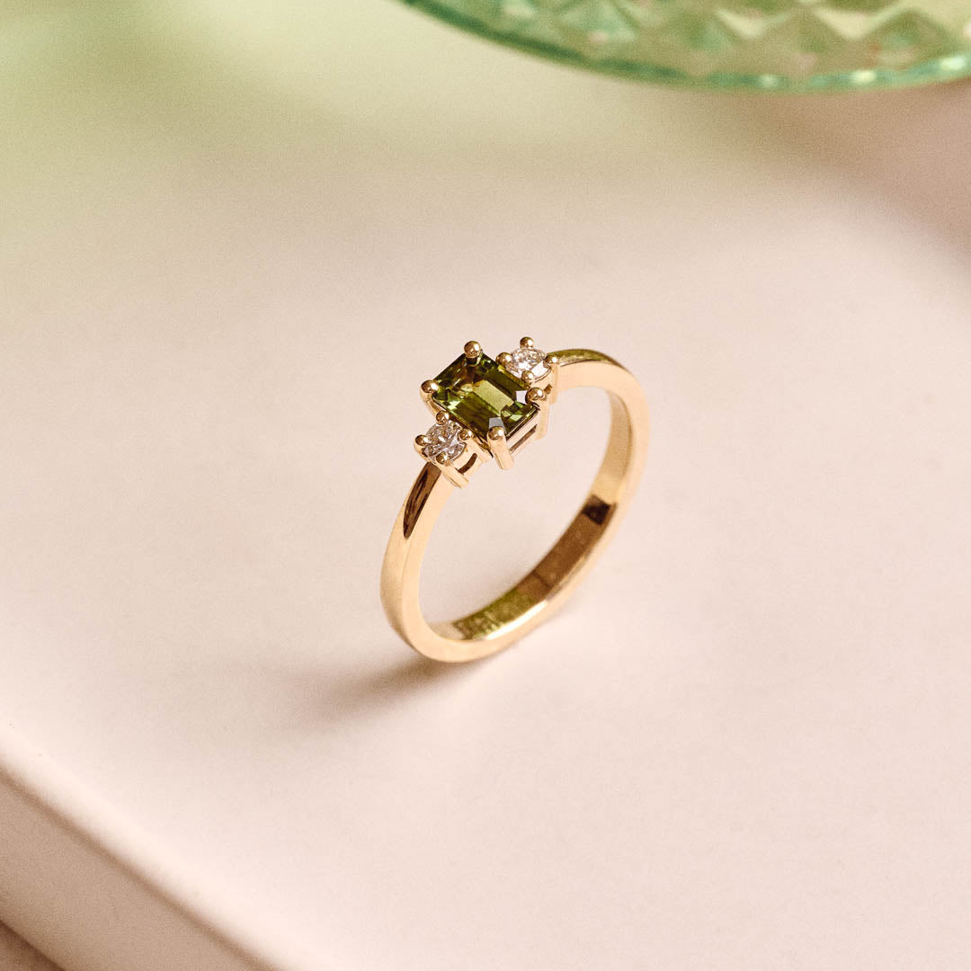 Handmade 9ct yellow gold trilogy ring featuring an emerald-cut green sapphire centre stone flanked by two round natural diamonds; elegant three-stone engagement ring design with timeless appeal.
