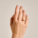 A close-up of a hand wearing a gold petite Botanica Protea Ring set with a champagne diamond, set against a solid light beige background.