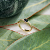Yellow gold handmade ring set with a central round 'peacock' sapphire and pavé diamonds down the shank on a green Protea leaf background