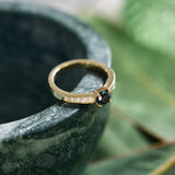 Yellow gold handmade ring set with a central round 'peacock' sapphire and pavé diamonds down the shank on a green blurred background