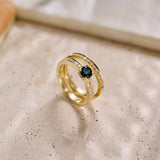 Yellow gold handmade ring set with a central round 'peacock' sapphire and pavé diamonds down the shank on a green Protea leaf background. Photographed with a yellow gold natural diamond eternity wedding ring on a textured background