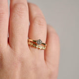 A close-up of a woman's hand wearing two stacked gold rings: one a solitaire round diamond ring in a Protea setting and the other with a delicate floral patterned curved wedding ring, photographed against a soft grey out of focus background.