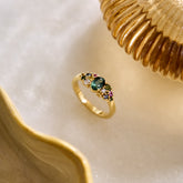 Yellow gold cluster ring set with a central teal sapphire and various additional coloured sapphires and natural diamonds on a white background with neutral tone accents