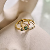 two yellow gold rings; one a cluster ring set with a central teal sapphire and various additional coloured sapphires and natural diamonds and the other a light blue sapphire eternity ring on a neutral textured background