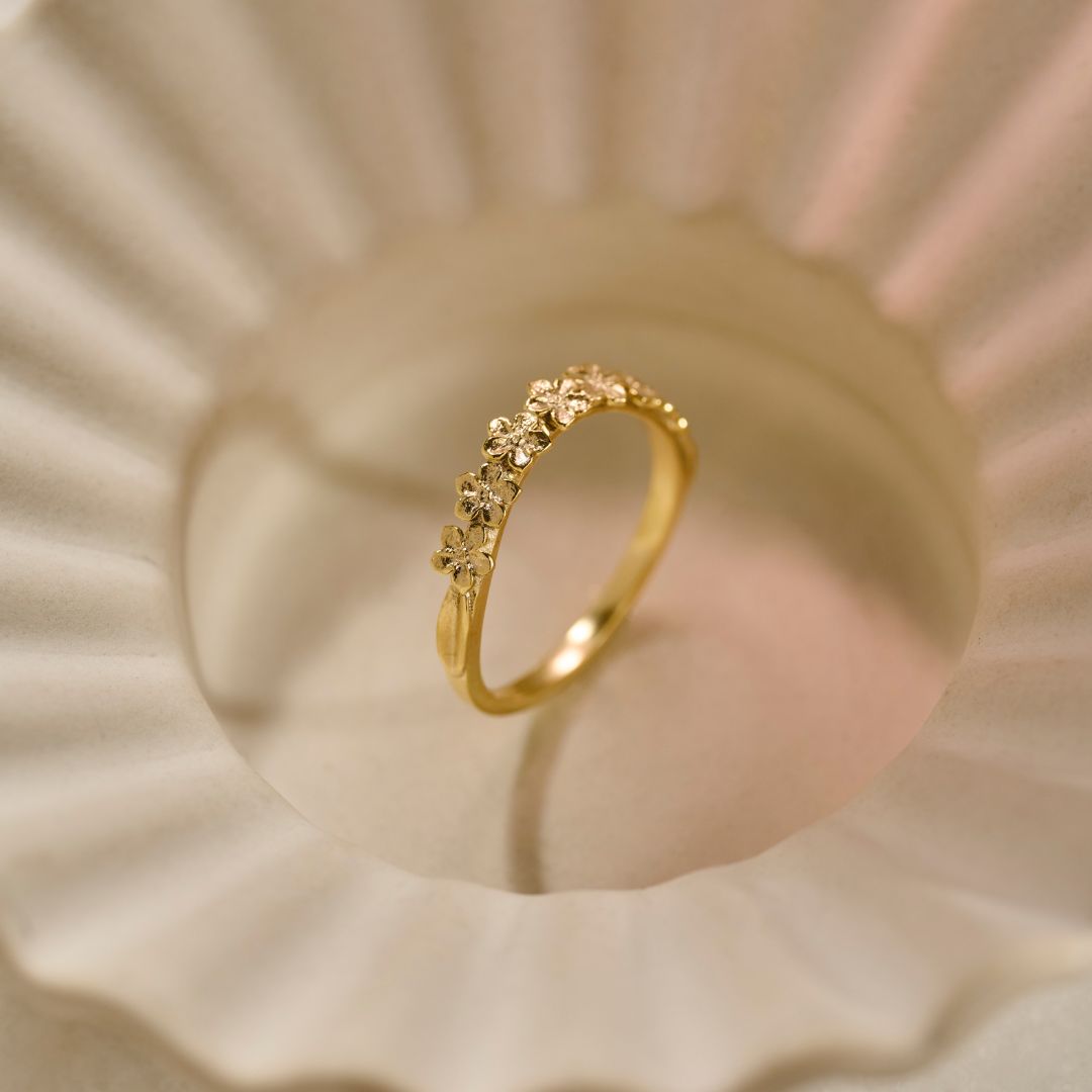 A delicately detailed gold wedding ring with a slightly curved row of small Forget-me-Not flowers, photographed on a textured, scalloped cream-coloured ceramic bowl