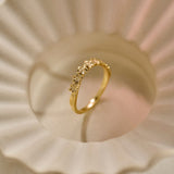 A delicately detailed gold wedding ring with a slightly curved row of small Forget-me-Not flowers, photographed on a textured, scalloped cream-coloured ceramic bowl