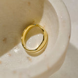 A polished yellow gold statement ring with an organic, wavy band shape photographed inside a textured cream-coloured stone dish.