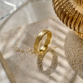 A textured organic yellow gold mens wedding ring positioned on a light natural stone slab with dappled shadows, alongside a decorative fluted glass candle holder in the upper right.