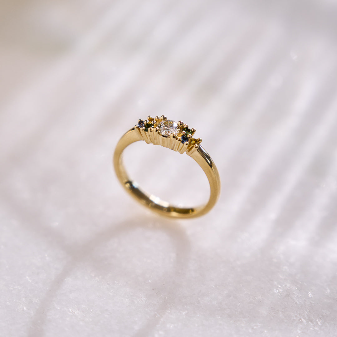 Yellow gold cluster ring set with a central natural diamond and additional blue, green, and yellow sapphires on either side. on a white background with subtle shadows.