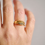 A close-up of a woman's hand wearing a gold ring featuring a cluster of natural diamonds and various coloured sapphires stacked with an organic yellow gold organic wedding ring, photographed against a soft light grey background.