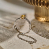Side view photograph of a oval yellow sapphire set in a four claw yellow gold setting and a platinum shank with pavé set round colour diamonds, resting on a textured beige stone surface.
