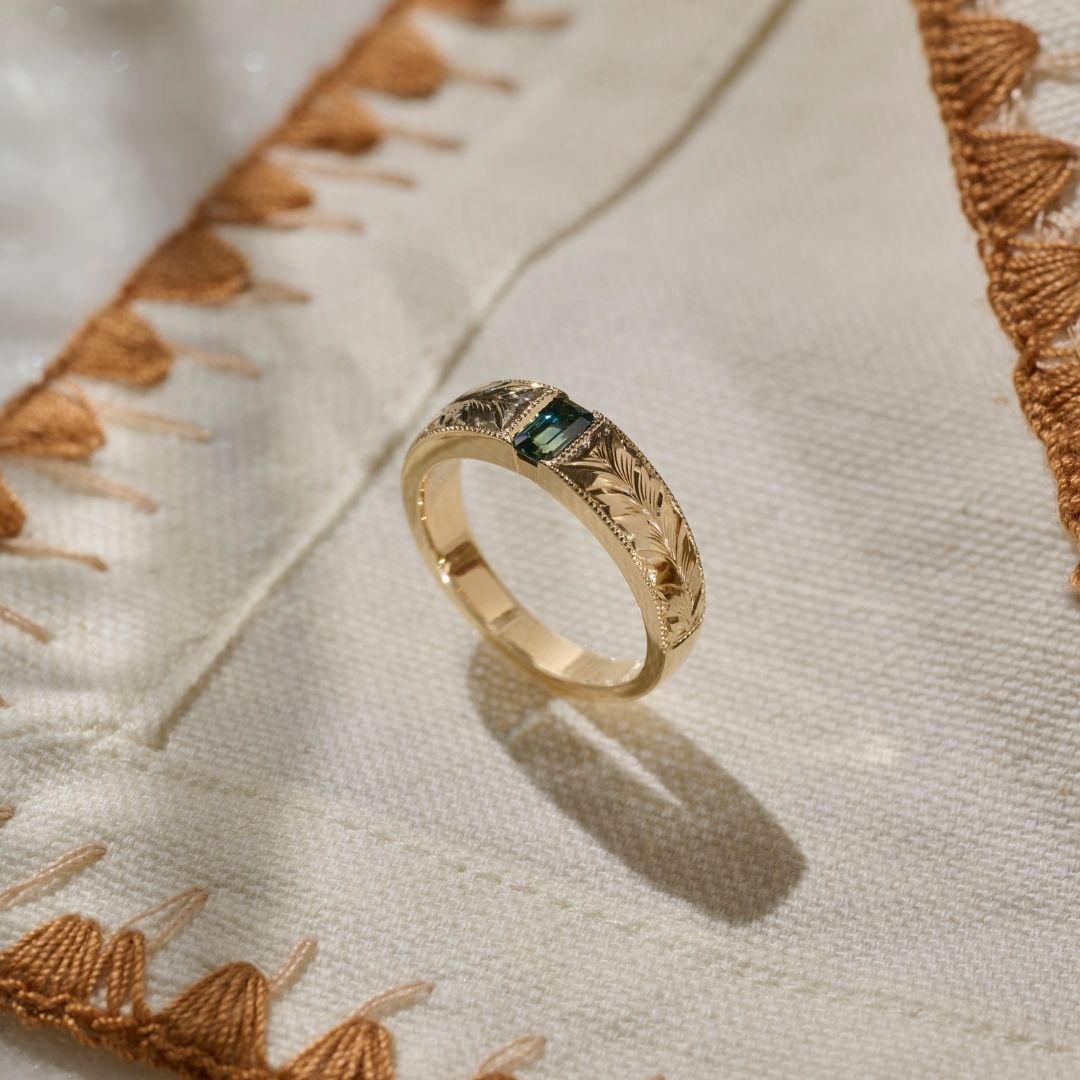 A hand made solid gold ring with an emerald cut green blue sapphire witha hand engraved leaf pattern, resting on a textured cream fabric. 