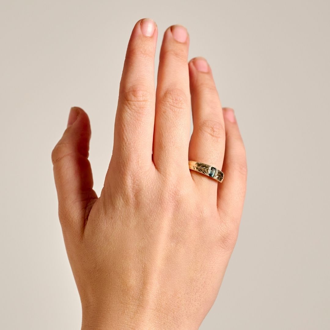 A close-up of a person's hand wearing a solid yellow gold ring showing its broad proportions on a hand and the hand engraved leaf pattern, set against a plain light gray background