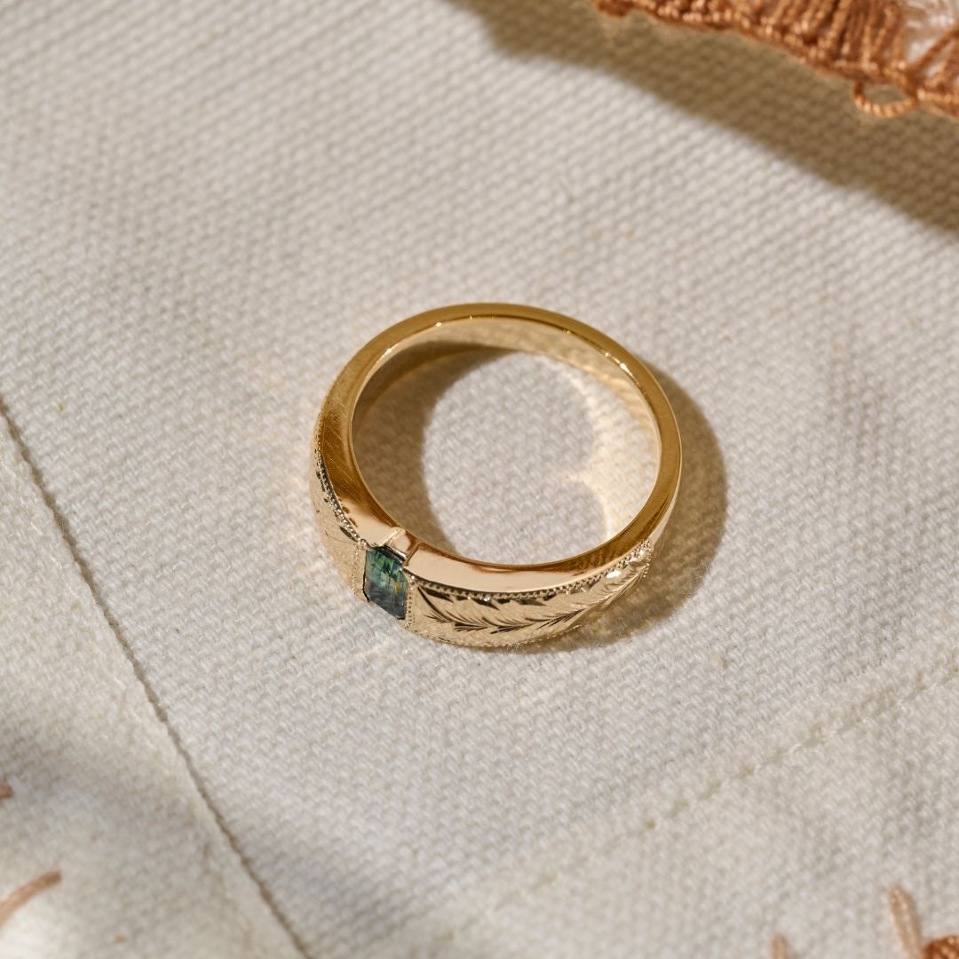 A solid gold ring with an emerald cut green  blue sapphire and hand engraved leaf detailing, resting on a textured cream fabric background.