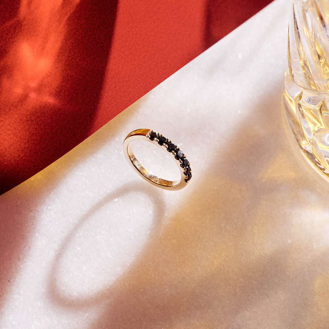 Handmade 9ct yellow gold wedding ring set with seven 2.5mm natural black diamonds; a modern, minimalist design perfect for unique wedding bands or alternative fine jewellery. Photographed on a red and white background and styled with a luxury whiskey glass.