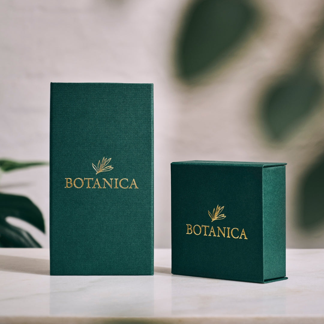 Two green 'BOTANICA' boxes on a light surface with a blurred green leafy background. Botanica Luxury Packaging.