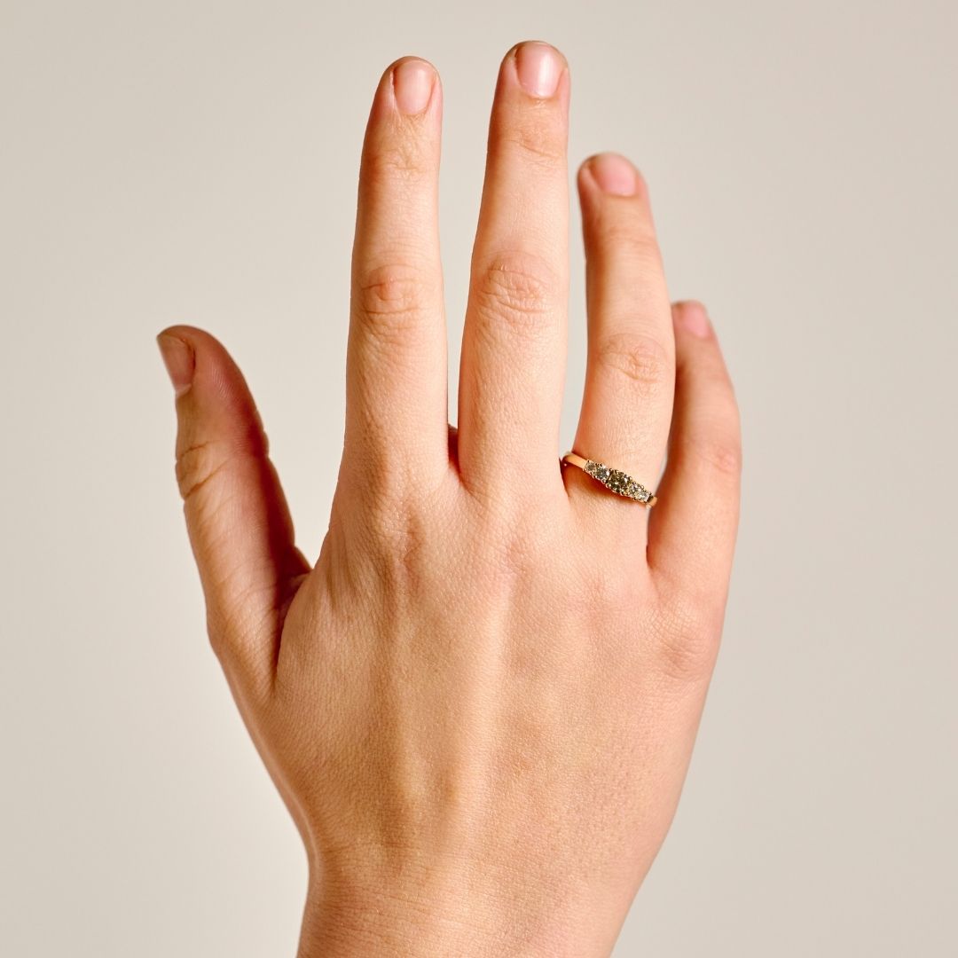 A close-up of a person's hand wearing the delicate solid gold five diamond with a central champagne diamond and four colourless diamonds flanking the central diamond, set against a plain light beige background.