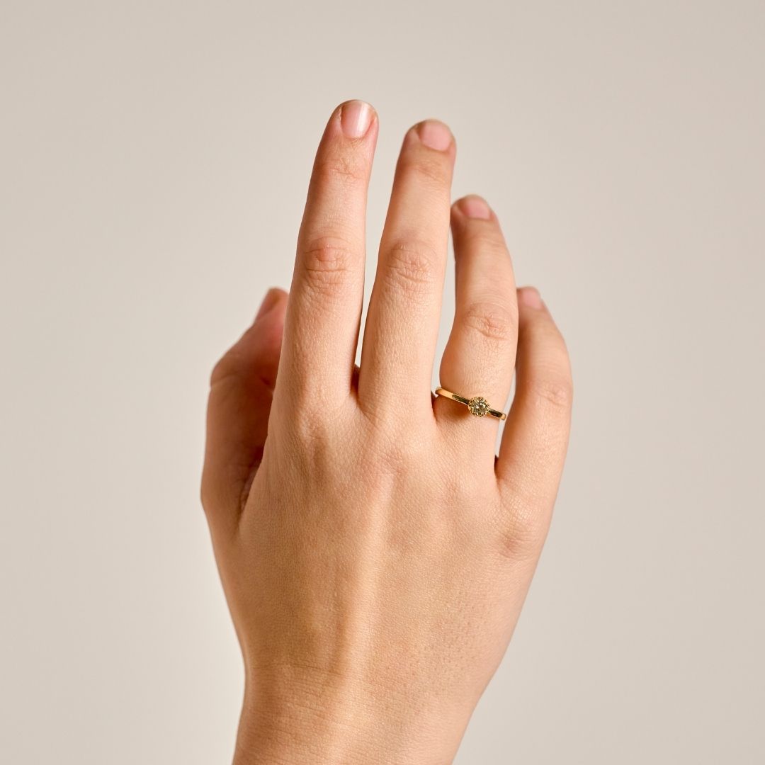 A close-up of a hand wearing a gold petite Botanica Protea Ring set with a champagne diamond, set against a solid light beige background.