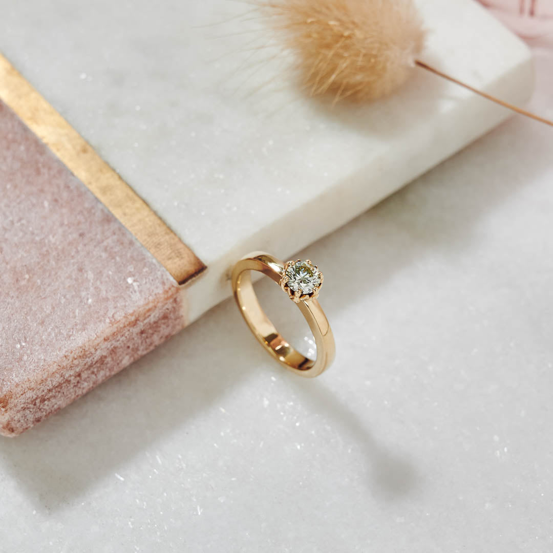 A solid 9ct yellow gold ring with a yellow toned diamond set in a protea setting, displayed on a marble surface with a soft focus background.