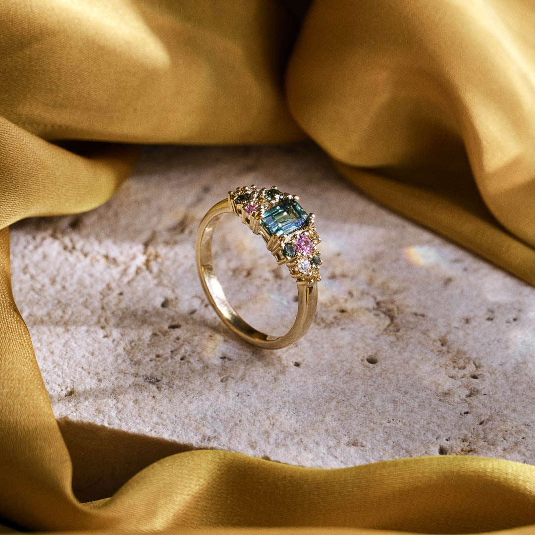 Handmade cluster engagement ring in 9ct yellow gold with a central emerald-cut green sapphire, surrounded by green, yellow, and pink sapphires and two natural diamonds in a floral-inspired design.