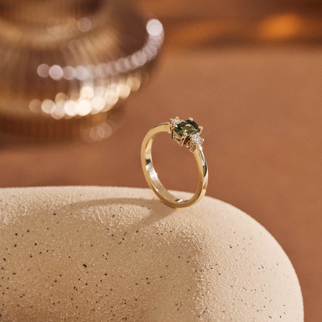 Handmade 9ct yellow gold trilogy ring featuring an emerald-cut green sapphire centre stone flanked by two round natural diamonds; elegant three-stone engagement ring design with timeless appeal.