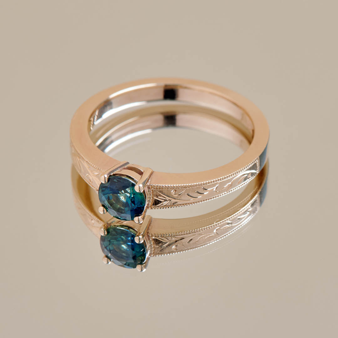A round teal sapphire set in a yellow gold ring with hand engraved botanical details on the band