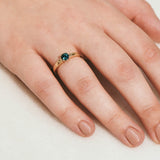 A round teal sapphire set in a yellow gold ring with hand engraved botanical details on the band - photographed on a hand.