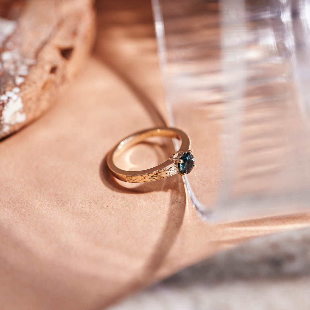 A round teal sapphire set in a yellow gold ring with hand engraved botanical details on the band