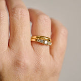 A close-up of a hand wearing the gold five diamond champagne ring, a central round fancy brown diamond flanked by four colourless diamonds tapering down in size row, paired with the path less taken wedding ring, a ring that is curved and organic, photographed against a soft light grey background.