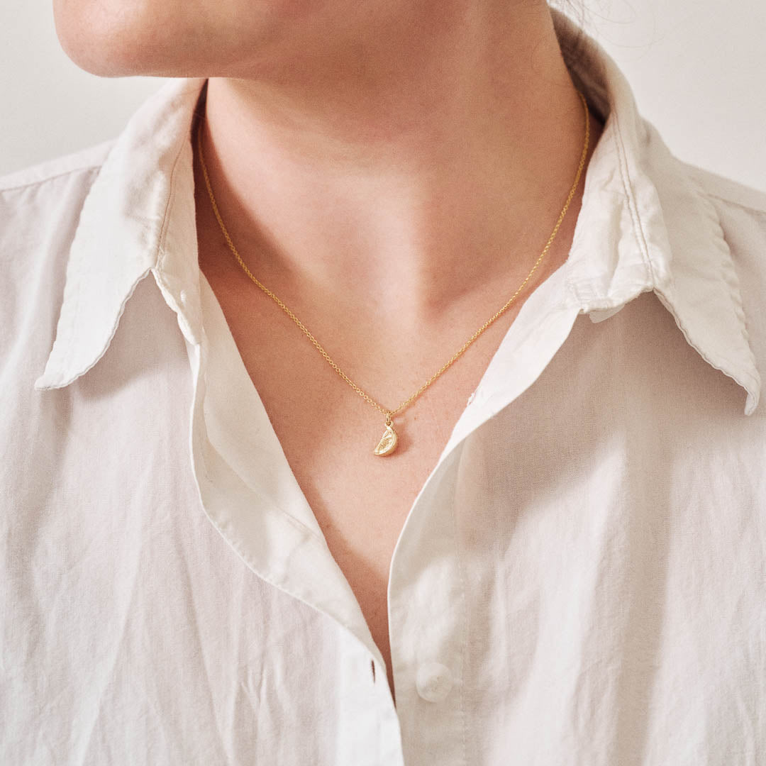 A miniature yellow gold citrus wedge jewellery charm on a yellow gold rolo chain. Photographed being worn.