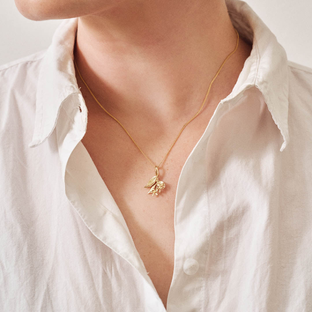 A yellow gold pendant featuring leaves and lemon blossom flowers on a yellow gold curb chain being worn.