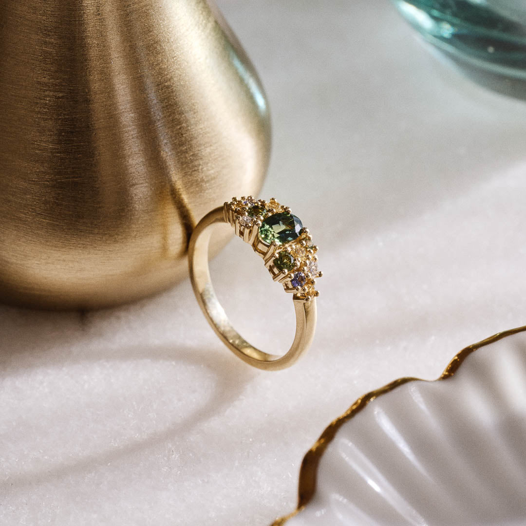 Unique handmade cluster engagement ring in 9ct yellow gold, featuring an oval green sapphire centre stone surrounded by a colourful bouquet-inspired mix of yellow, green, and blue sapphires with two natural diamonds.