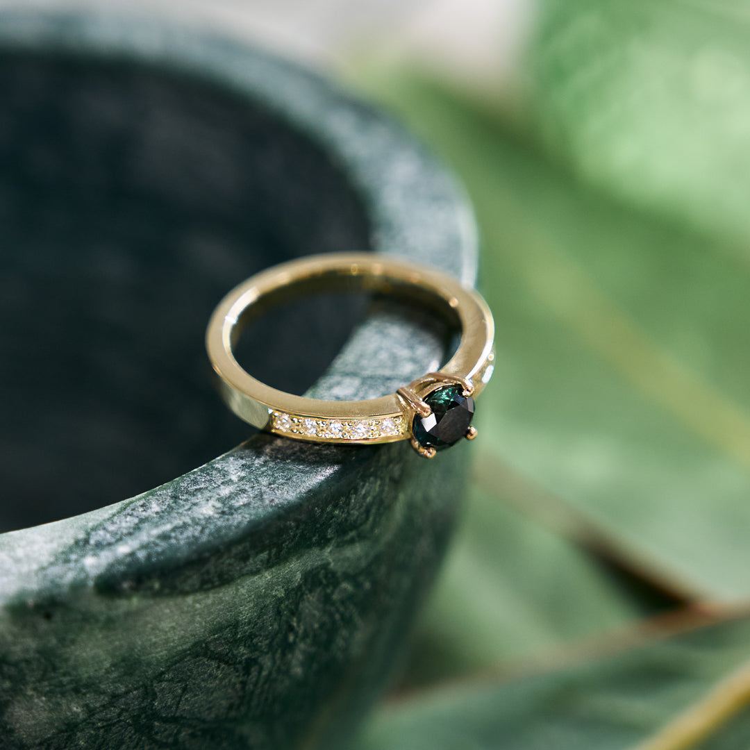Yellow gold handmade ring set with a central round 'peacock' sapphire and pavé diamonds down the shank on a green blurred background