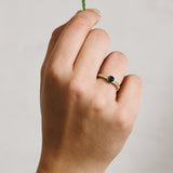Hand wearing a yellow gold ring with a green-blue sapphire and natural diamonds on a neutral background
