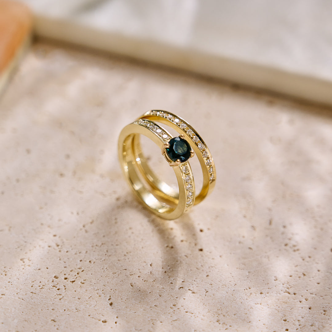 Yellow gold handmade ring set with a central round 'peacock' sapphire and pavé diamonds down the shank on a green Protea leaf background. Photographed with a yellow gold natural diamond eternity wedding ring on a textured background