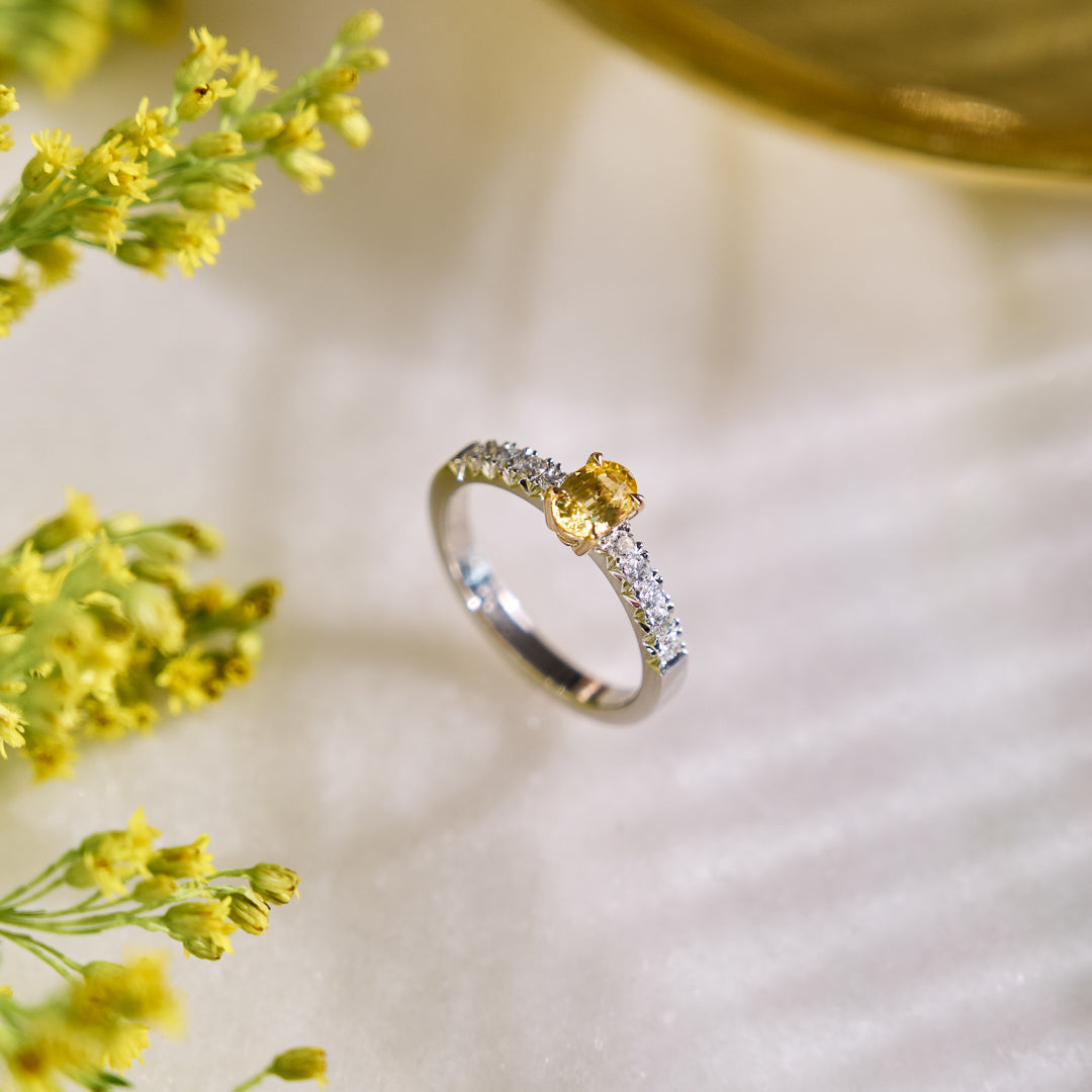 Platinum ring with central yellow sapphire set in yellow gold, and french pave natural diamonds. Surrounded by yellow flowers on a light background