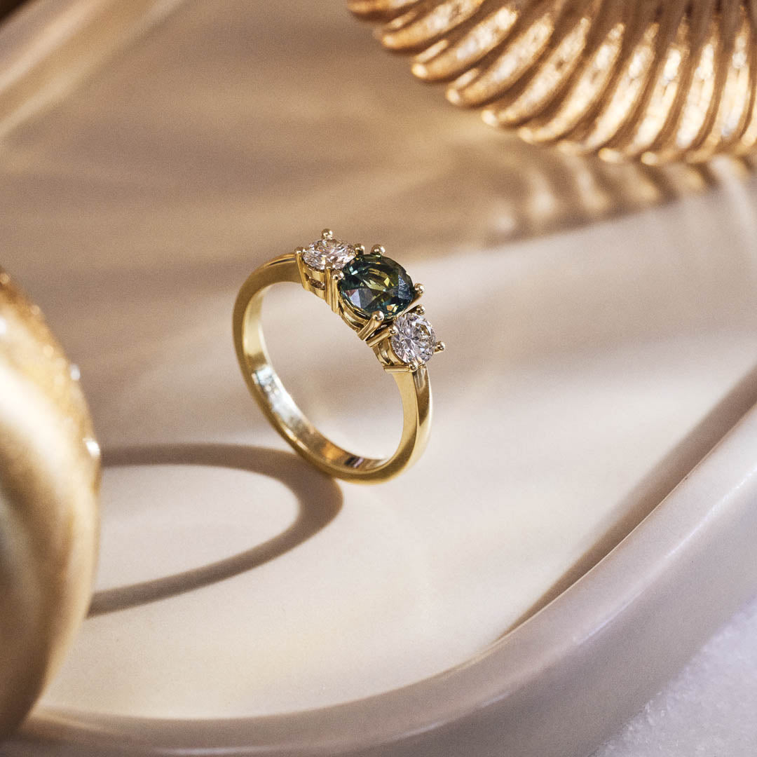 Handmade 14ct yellow gold engagement ring featuring a round green sapphire centre stone flanked by two natural round diamonds; luxury heirloom-quality design perfect for weddings or special occasions. Styled and photographed on a grey stone soap dish with fluted glass details and moody lighting.