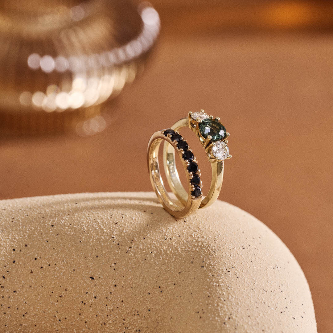 Handmade 14ct yellow gold engagement ring featuring a round green sapphire centre stone flanked by two natural round diamonds; photographed with Botanica's Black Diamond and 9ct Yellow Gold wedding ring. Photographed on a textured vase with a terracotta coloured background and a fluted glass detail blurred in the background.