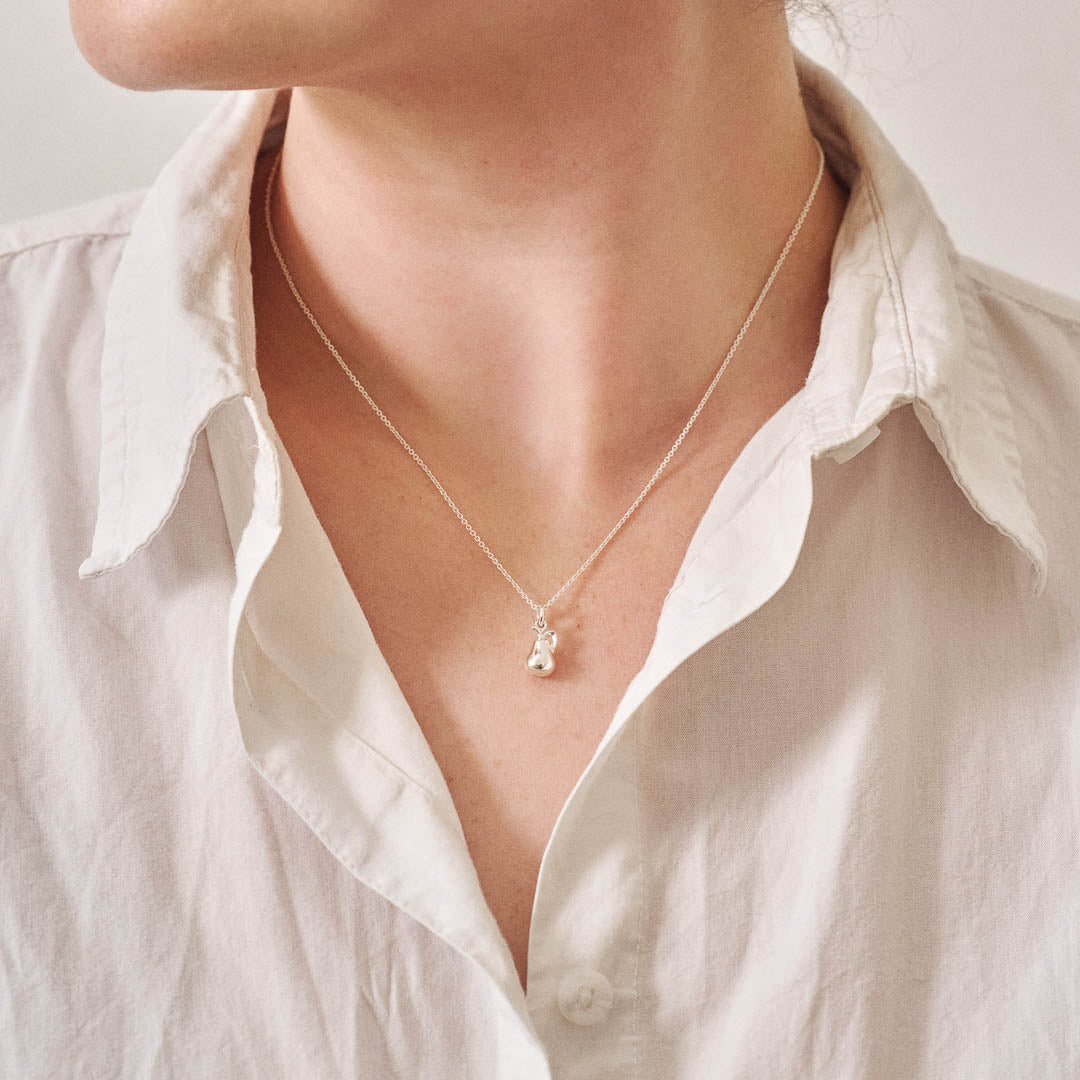 A miniature pear jewellery charm in silver, on a silver rolo chain, photographed being worn.