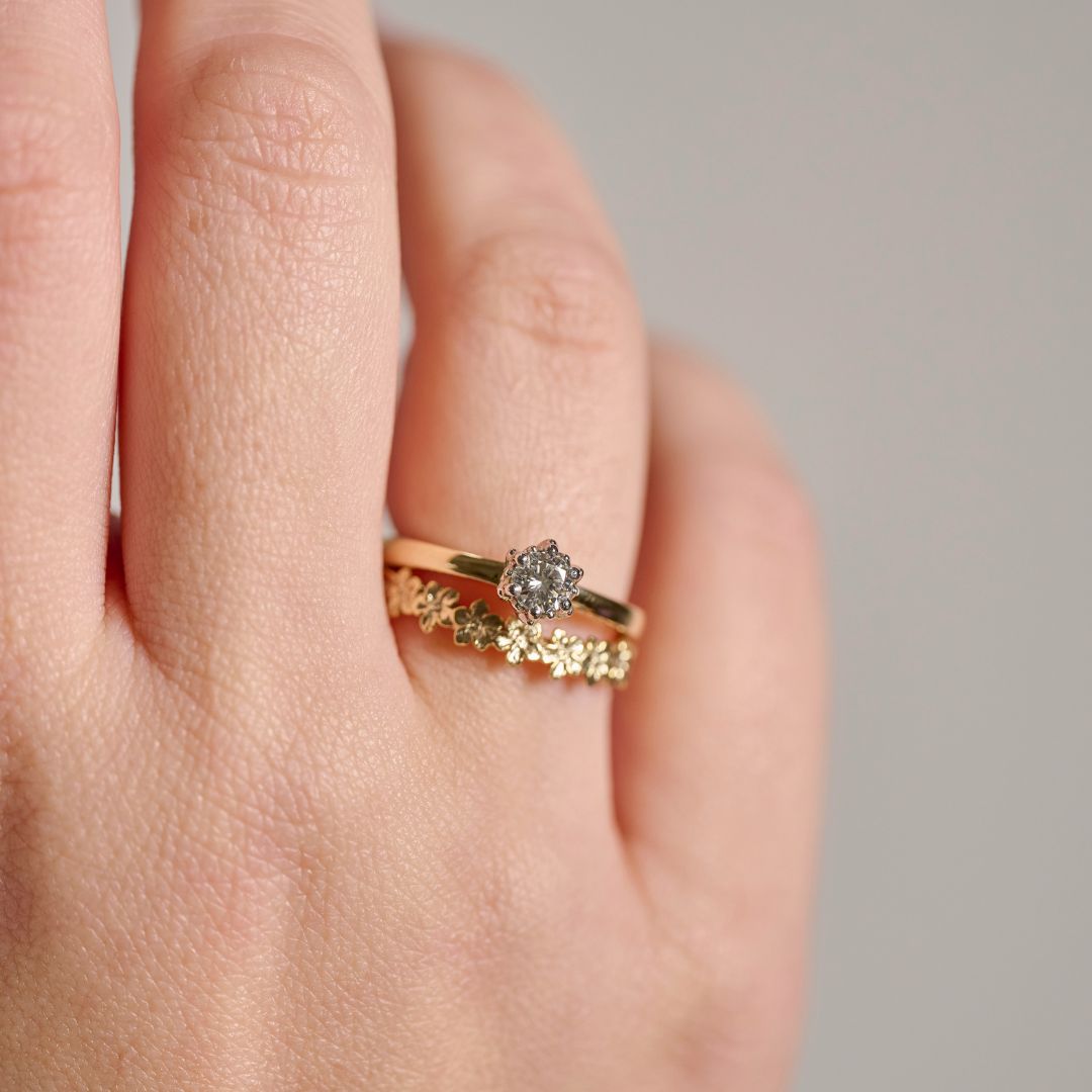 A close-up of a woman's hand wearing two stacked gold rings: one a solitaire round diamond ring in a Protea setting and the other with a delicate floral patterned curved wedding ring, photographed against a soft grey out of focus background.