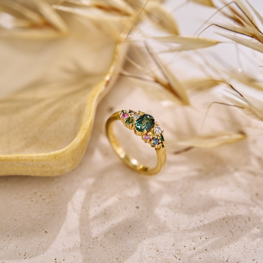 Yellow gold cluster ring set with a central teal sapphire and various additional coloured sapphires and natural diamonds on a neutral textured background with dried grasses