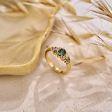 Yellow gold cluster ring set with a central teal sapphire and various additional coloured sapphires and natural diamonds on a neutral textured background with dried grasses