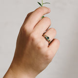 Hand wearing a yellow gold cluster ring set with a central teal sapphire and various additional coloured sapphires and natural diamonds on a white background