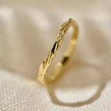 A close-up photograph of a 9ct yellow gold wedding ring with a textured hand carved leaf pattern, resting on an out of focus textured cream fabric background.