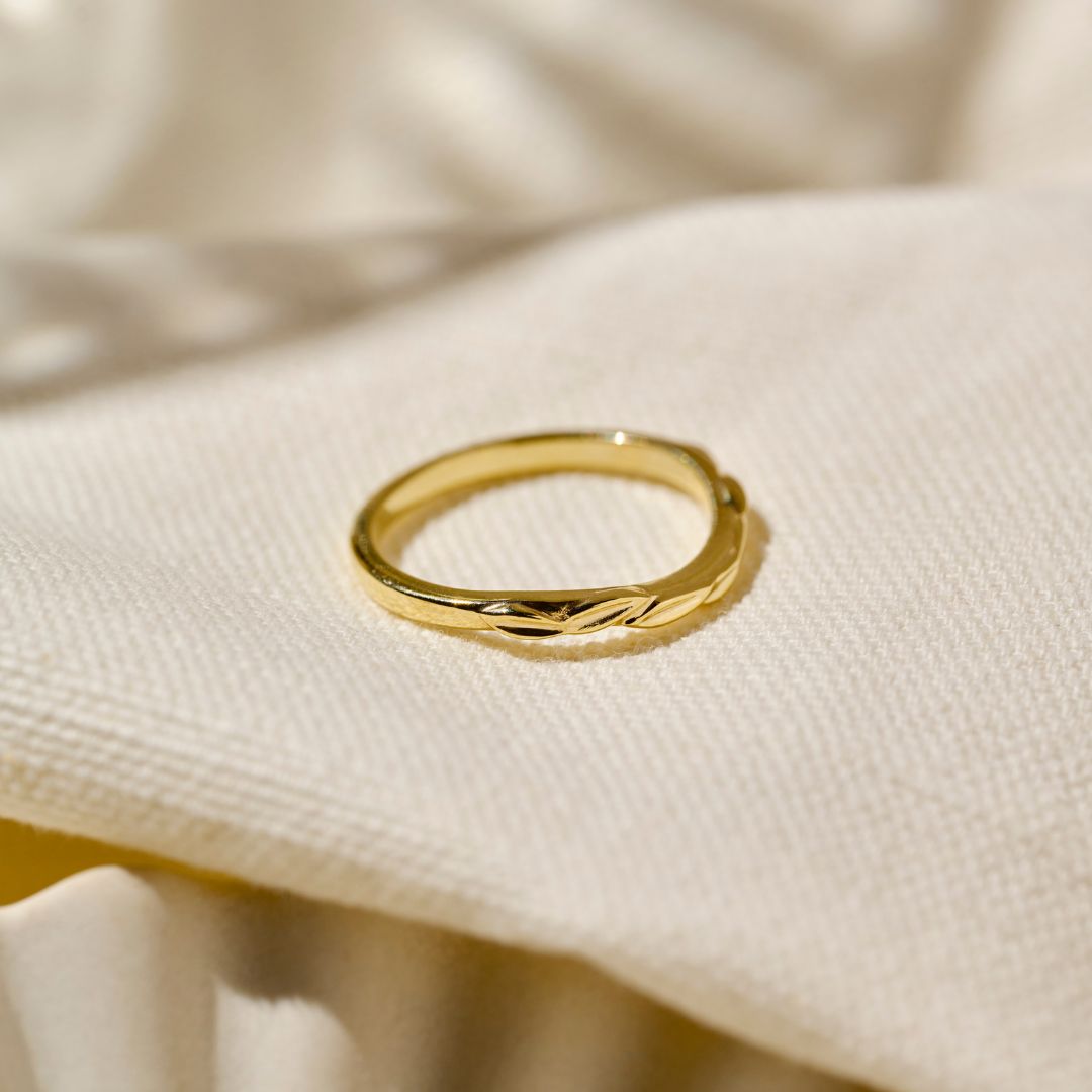 A leaf inspired unique 9ct yellow gold wedding ring positioned on its side with a wavy hand carved leaf design, resting on a textured cream-coloured fabric background.
