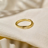 A leaf inspired unique 9ct yellow gold wedding ring positioned on its side with a wavy hand carved leaf design, resting on a textured cream-coloured fabric background.